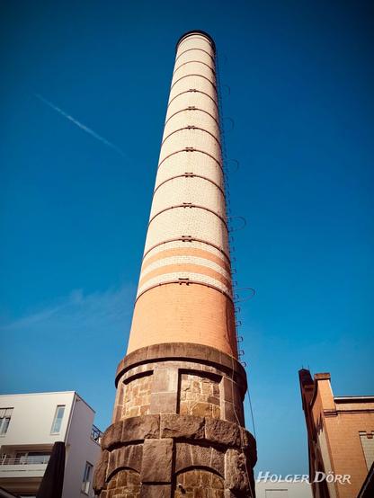 Ein historischer Industrieschornstein des „Alten Schlachthofs“ in Gießen ragt in den strahlend blauen Himmel. Der Schornstein ist aus Backstein gebaut, mit hellen und dunklen Ziegelabschnitten sowie metallischen Verstärkungsringen. Eine rostige Leiter führt spiralförmig an seiner Seite hinauf. Der massive Sockel besteht aus grob behauenen Natursteinen und gemauerten Elementen. Rechts im Bild ist ein Teil des alten Backsteingebäudes zu sehen, links ein modernes weißes Wohngebäude. Die Perspektive von unten verstärkt die beeindruckende Höhe des Schornsteins.