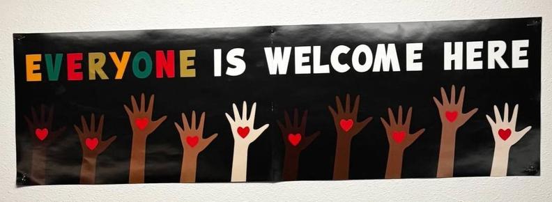 Sign saying "Everyone is welcome here" is all capital letters. Beneath the sign is a series of hands of various colors. Each hand has a red heart on it.