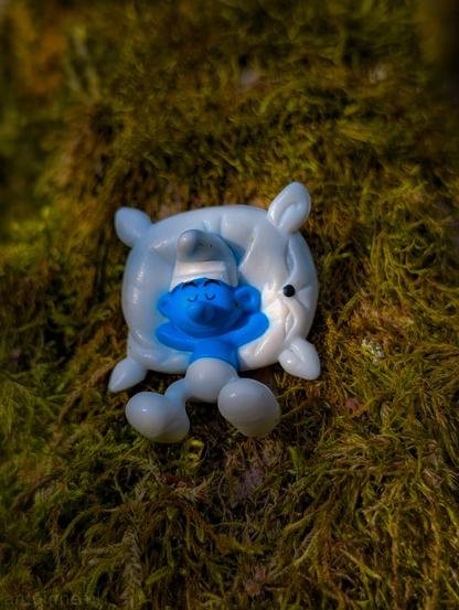 A smurf lying on its back with its eyes closed. He is resting on a small, fluffy-looking white pillow, positioned on a patch of moss.