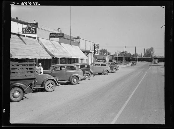 This black and white photograph depicts a bustling main street in Nyssa, Oregon during what appears to be the 1930s. Several vintage cars are parked on both sides of the road alongside trucks loaded with goods. Visible businesses include "Meats," which suggests butchery services or possibly sales, as well as establishments like Texaco service station and a John Deere dealership that indicates availability for agricultural equipment maintenance and purchases.

The architecture is modest yet functional, reflecting an era where commerce was likely local-focused without the presence of modern skyscrapers. A few pedestrians are visible on foot in front of one truck with wooden crates loaded onto it. The sky above suggests clear weather conditions during this time period.