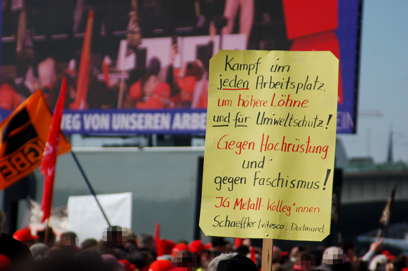Pappschild auf der Kundgebung der IG Metall: "Kampf um jeden Arbeitsplatz, um höhere Löhne und für Umweltschutz! Gegen Hochrüstung und gegen Faschismus! (IG Metall Kolleg*innen Schaeffler / vitesco, Dortmund). Im Hintergrund rote Gewerkschaftsfahnen vor dem Großbildschrim an der Bühne