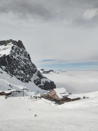 Foto. Berglandschaft mit Schnee, Nebelmeer, Gebäude und Sesselbahn.
