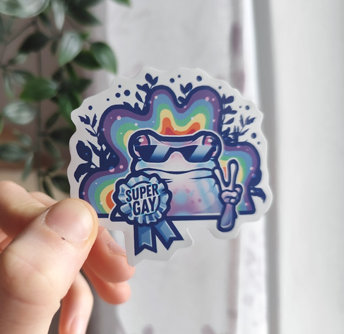 Hand holding up a sticker of a colorful frog with glasses and a wavy rainbow behind it, showing a peace sign and rocking a "super gay" award ribbon.