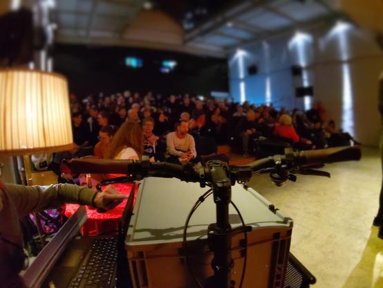 Eine graue Kunstoffbox mit Schublade steht auf der Ladefläche eines Lastenrads. Auf der Schublade steht ein Laptop, der von einer Person bedient wird. Im Hintergrund unscharf das Publikum im vollbesetzten Kino
