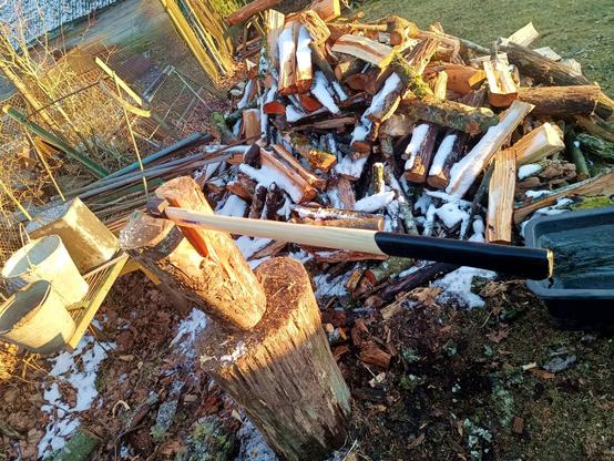 A new axe, stopped in a log that it has split but hangs on with a few fibers on top of a chopping block. A pile of slightly snowy firewood behind, with some freshly split logs on top.