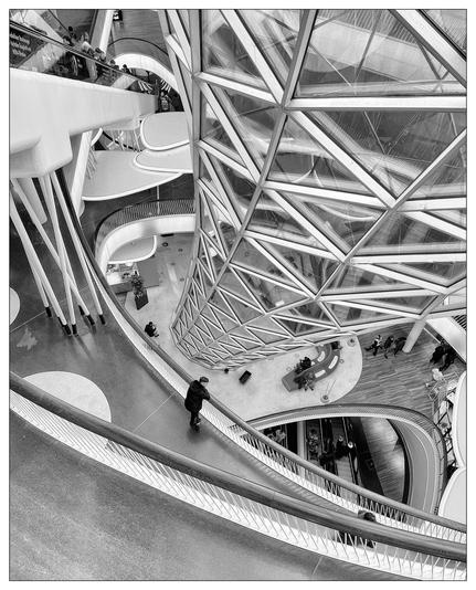 The image presents a modern architectural interior of a shopping center. The photo captures multiple levels connected by sleek, winding staircases and escalators. The prominent feature is a geometric, glass-paneled structure that forms part of the ceiling or façade, allowing natural light to flood the space. This complex lattice of steel and glass contrasts with the smooth curves of the staircases and the flat, clean lines of the railings and floors. People can be seen moving through the space or standing on different levels, adding a human element to the scene. The image is black and white, which emphasizes the patterns, shapes, and light contrasts within the architectural design.