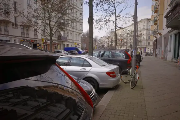 A row of parked cars next to a sidewalk with a lamp pole which has an old bike attached to it. Parts of the buildings on the left and right side of the street are visible with a few shops and restaurants.