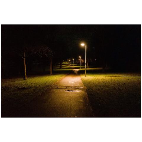 A colour photo at night. In the middle is a pedestrian way well lit by street lamps. To the left and right is mostly grass and some trees. The circles of the light from the street lamps and the colour temperature of the light give the photo its style.