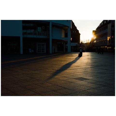 A colour photo on the minster place in Ulm. On the left and middle is the Stadthaus, on the right edge another building. Between is a gap between the buildings giving way to the descending sun which gives the whole scenery a rather warm colour. A person is standing in the middle of the beam of light and throwing a long shadow.