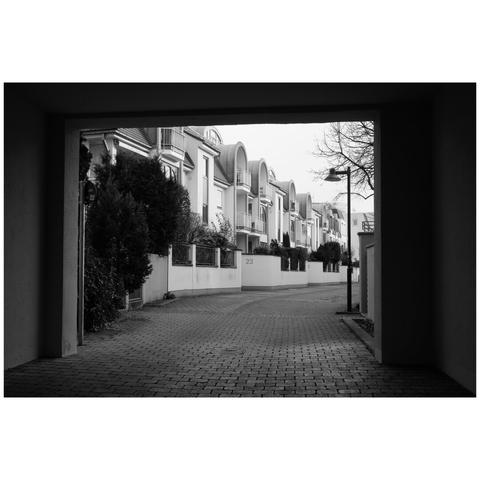 Black and white photo. The scene is framed by a rectangular archway. A row of of buildings all of the same architecture can be seen from the middle of the frame going towards the background. The buildings all have a small frontyard surrounded with brick walls.