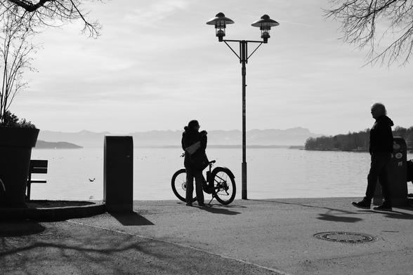 A black and white photograph captures a lakeside scene at Lake Starnberg, Bavaria, with silhouettes of people, a bicycle, and a lamppost against a calm water and mountain backdrop. A woman with a bicycle stands near the water, while an older man walks past. The lamppost, trash bin, and bench add depth to the composition. The soft sky and distant mountains contrast with the dark foreground elements.