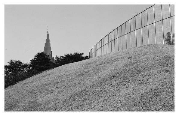 Black and white photograph in Shinjuku Gyoen Park. You can see a meadow hill in the foreground, on which the glass front of the greenhouse rises and stretches from the right to the center of the picture. The curves of the hill and the glass front are smooth and almost glide into each other. A few trees can be seen in the left part of the picture and a high-rise building in the background.
