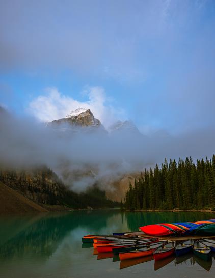 Colourful moored canoes reflected in the calm turquoise waters of Moraine Lake with snow peaked mountains peeking above low fog in the background, Banff National Park, Alberta, Canada
Captured by Komeil Karimi