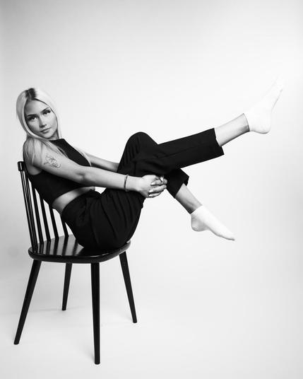 The image shows a black and white photograph of a person sitting sideways on a chair. They are wearing dark pants, a sleeveless top, and white socks. They have one leg extended and are holding their leg with both hands. The person has long hair and is looking at the camera.