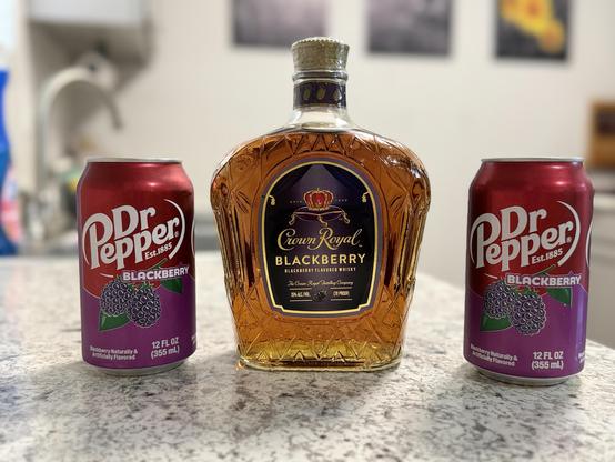 The image features a bottle of Crown Royal Blackberry Flavored Whisky centrally placed on a countertop, flanked by two cans of Dr Pepper Blackberry soda. The whisky bottle has a distinctive, ornate shape with a gold-colored cap and a label that prominently displays the Crown Royal logo and the words "Blackberry Flavored Whisky." The whisky is amber-colored, indicating its alcohol content. The Dr Pepper cans are red with a purple label, featuring the brand's logo and the words "Blackberry" and "Est. 1885." Each can is labeled as containing 12 fluid ounces (355 mL) of blackberry-flavored soda. The background is slightly blurred, showing a kitchen setting with a sink and some wall decorations.