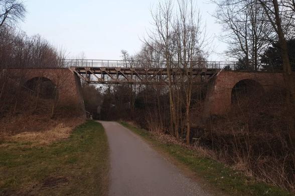 Verstrebte Stahlbrücke zwischen Gemäuer aus roten Ziegeln in rötlichem Abendlicht. In Bildmitte breiter Fußweg, rechts der breite, ruhige Bach. Bäume und Sträucher laublos. Der Boden voller Laub: grau-braun. Grünes Gras am Wegesrand.