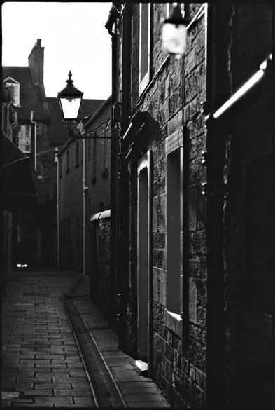 Black and white photography of a narrow street in opposing light.
An old-style street lamp draws the focus.