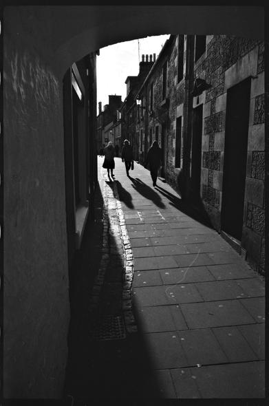 Black and white photography of a narrow street in opposing light.
Several people are creating a strong shadow.
