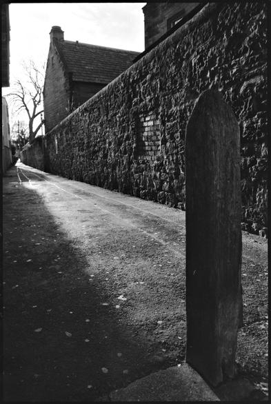 Black and white photography of a narrow street in opposing light.
A bollard is very prominent in the foreground.