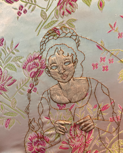 An embroidered portrait of a woman with the face and neck mostly filled in a pale skin tone in satin and long and short stitch’s