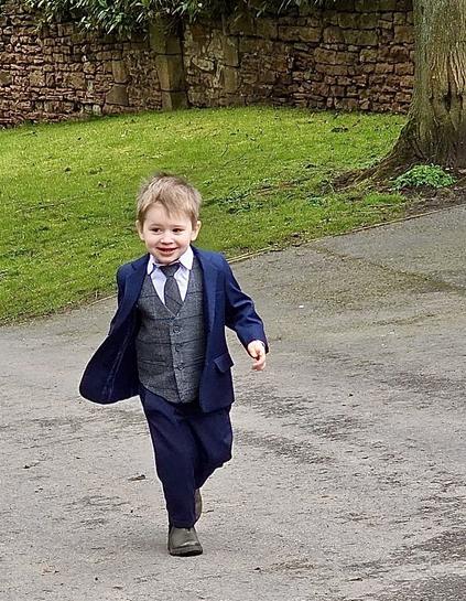 A young boy in a formal outfit runs on a pathway, smiling. He wears a dark blue suit with a gray vest and a white shirt, surrounded by greenery and stone walls in the background.