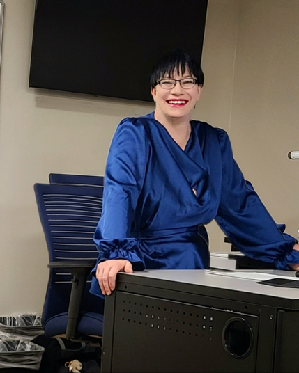 A smiling, dark-haired person leaning on a desk. They're wearing a blue satin top and rectangular eyeglasses.