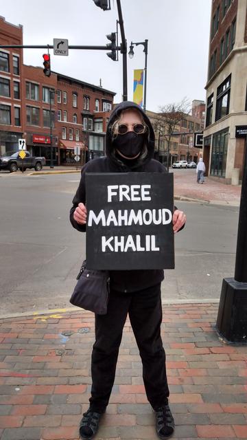 A person on a street corner, wearing black, and holding a sign that reads "FREE MAHMOUD KHALIL".