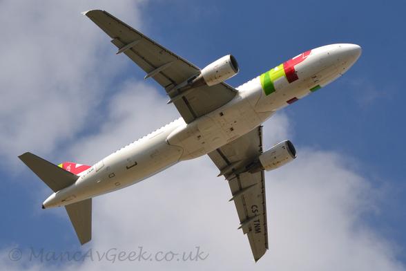 View from underneath of a white, twin engined jet airliner flying from left to right almost directly overhead at a very low altitude, with undercarriage tucked away and flaps extended from the rear of the wings, suggesting it has just taken off.
There are large, billboard-style letters "TAP" in dark green, light green, and red, respectively, on the forward fuselage, a design replicated at least in part on the tail.
The registration "CS-TNM" is on the lower rear fuselage and under the left wing in black.
Behind it, the sky is a mix of pale blue, and fluffy white.