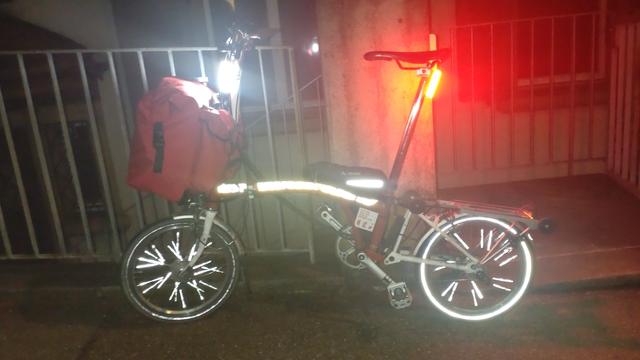 Bild eines rot-weissen Brompton-Faltrades mit gefüllter roter Fronttasche an ein Metallgeländer gelehnt. Ei ist dunkel, an der Lenkstange und Sattelstange sind zwei sehr helle 270 Grad LED-Leuchten montiert und an. Das Bild ist mit Blitz aufgenommen und es reflektieren in weiss die obere Hälfte des Hauptrohres des Rahmens, ein Streifen auf der kleinen Tasche auf dem Hauptrohr, stäbchenförmige Reflektoren auf den Speichen sowie reflektierende Streifen auf den Reifen: Hinten sehr gut und deutlich, vorne nur teilweise und schwach.