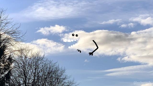Photo of a blue sky with a few fluffy white clouds, one big solid one near the centre and other more wispy ones above and to the left. Some large bare tree tops cut across the bottom left corner of the frame. The main cloud has a v-shape cut out to which I have added large flat teeth so it forms a chompy jaw. I have also added eyes above, and arms.