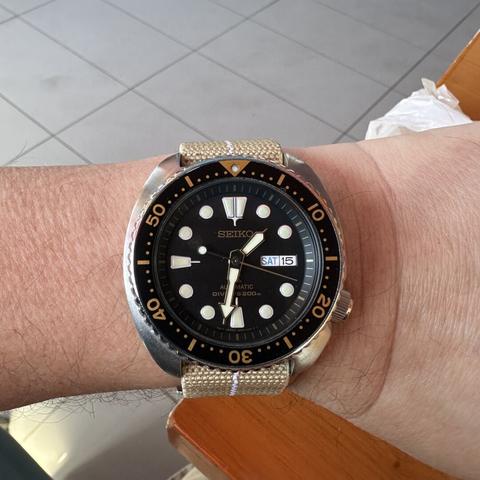 A watch with brushed stainless steel cushion case and 4:00 crown with black gilt dial, 3:00 day/date, gold hands with white lume, and black dive bezel with gold markings, on a khaki elastic strap with white centerline.