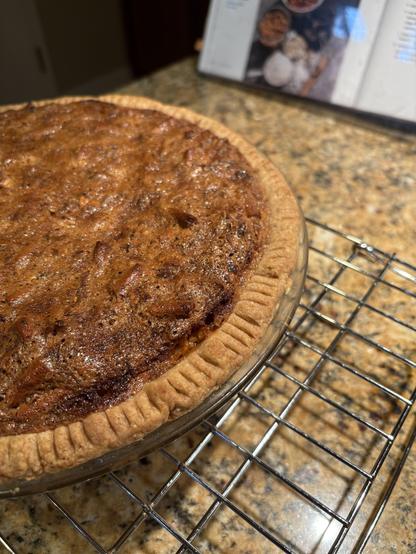 Close-up of a just-baked pie on a cooling rack, with cookbook visible in background. The pie crust is dark golden (partly whole wheat), decorated with fork tine impressions around the edge. The filling is dark golden brown with even darker points here and there where a piece of pretzel (substituted for nuts) pokes through the caramelized crust of the surface.