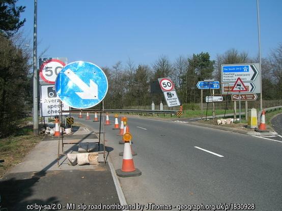 A view along a 2-lane sliproad, with trees in the distance.  Half the width of the road is closed with orange traffic cones, and a large portable "keep right" sign (a blue circle with a white arrow) dominates the foreground. Elsewhere are a jumble of permeant direction signs and temporary speed limit signs.