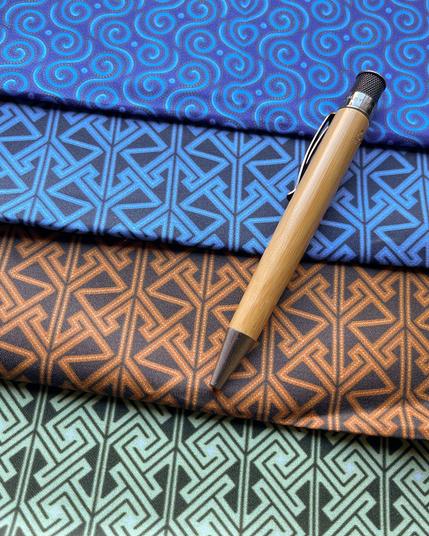 Four pieces of fabric arranged like shingles: a field of spirals in shades of blue, a Celtic key / maze pattern in an almost-black on blue, the same key / maze pattern on a warm brown, a different key /maze pattern (like squared spirals) in dark on a minty green.  A bamboo body pen sits at an angle across the stack (for scale).
