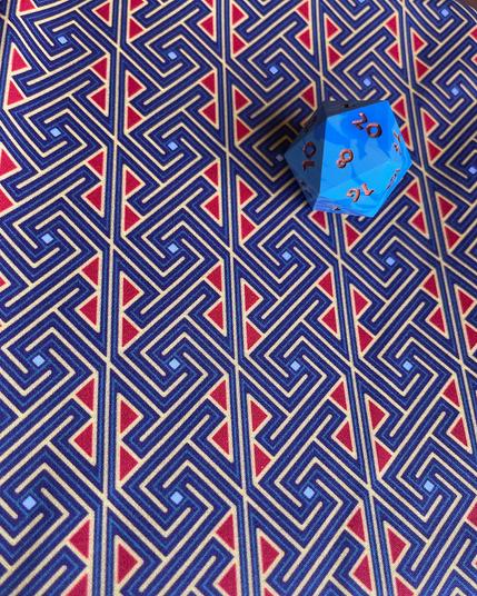 A piece of fabric adorned with a Celtic key / maze pattern that looks kind of like spirals but square in blues, red, and gold lying flat with a marbled blue 20-sided die with metallic copper numbers.