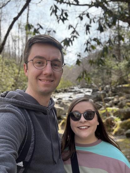 A smiling couple taking a selfie outdoors in a wooded area with a small rocky stream in the background. The man is wearing glasses, a gray hoodie, and a backpack, while the woman is wearing sunglasses and a colorful striped sweater. Sunlight filters through the trees, casting natural light on their faces.
