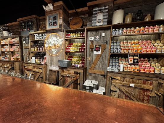 The interior of a rustic moonshine distillery store Ole Smoky, with wooden shelves lined with colorful jars of moonshine. A brown and white neon sign reads “Ole Smoky Holler,” and various decorations, including vintage jugs and signs, are displayed on the walls. A copper bar counter sits in the foreground for a round of moonshine tasting.