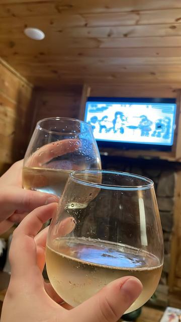 Two people hold out wine glasses with a standard pour of champagne in each inside of a natural wooden interior. A TV is playing the movie Diary of a Wimpy Kid: Rodrick Rules.