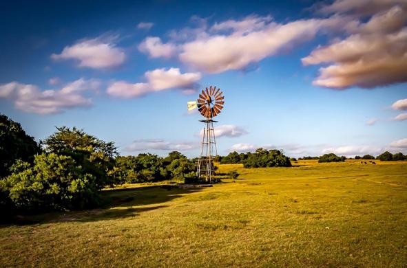 The image shows a rural landscape with a windmill on a grassy field. The sky is blue with scattered clouds, and there are some trees around the field. The scene has a peaceful and open countryside feel.