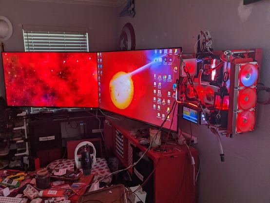 Photo of a red PC mounted on a wall next to two large TVs. The PC components are mostly red and illuminated by red and white lights. It is cooled by a liquid cooling system pumping red fluid through clear tubing.
