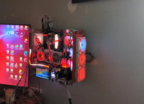 Photo of a red PC mounted on a wall next to a TV. The PC components are mostly red and illuminated by red and white lights. It is cooled by a liquid cooling system pumping red fluid through clear tubing.