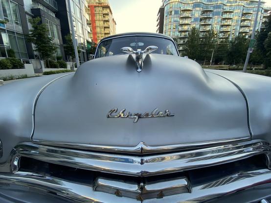 Front grille of an old car (possibly early-1950s), with a black roof and silvery-pearl body. We are so close to it the headlights are outside the frame’s sides, and you can barely see the roof.