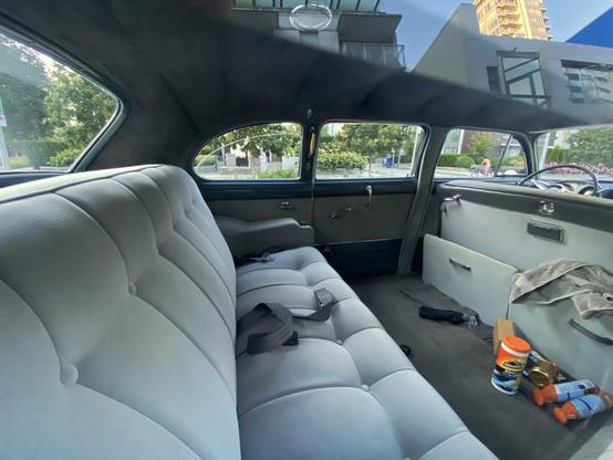 Interior, rear passenger area of an old car (possibly early-1950s). There is a massive tear bench seat, and enough floor space to allow a fully grown person to lie down on without fear of their shoulders being cramped.