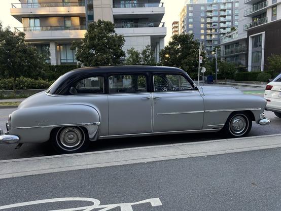 Very old car (possibly early-1950s), with a black roof and silvery-pearl body, seen in full side angle.