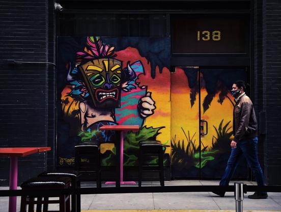 A person with a mask walks past a closed bar with a wall mural decorated with bright colors in a tiki theme
