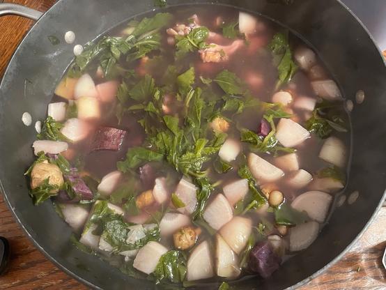 A pot filled with soup containing chopped vegetables, including leafy greens, various root vegetables, and a clear broth.