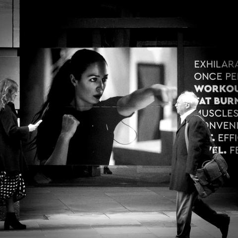 Black and white. An older man is walking into the picture from the right, and a young woman from the left. Behind them both, in the middle of the image is a five feet high poster featuring a photo of a young woman practising some form of boxing without gloves. She is facing to our right and punching with her left hand, and her fist is about to come out of the poster and strike the man's face.