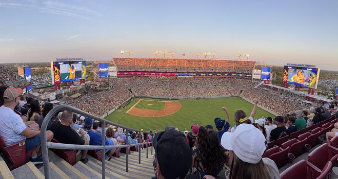 A large crowd fills Raymond James Stadium, which has been temporarily converted into a baseball field. The stadium is packed with fans, with the seating area illuminated by the soft glow of the setting sun. The field has a dirt infield and lush green grass in the outfield. Players are scattered across the field, with one on the pitcher’s mound. A digital scoreboard and advertisements are visible on the left side of the image, including a sign that says “Go Bucs,” indicating the stadium’s usual purpose as a football venue. Fans in the foreground are watching the game from high up in the stands.