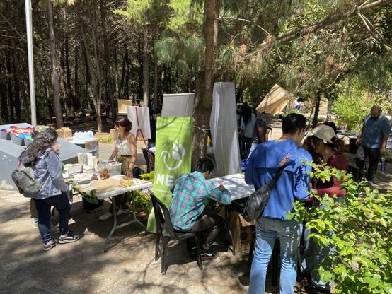 La fotografía muestra en primer plano a una chica haciendo una presentación a dos personas, en un parque público. Alrededor de la mesa en donde está ella hay un grupo de personas realizando actividades artísticas