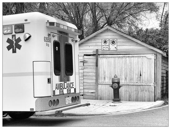 This black and white image features the rear of an ambulance parked on a street corner. The ambulance has the universal medical emblem (a star of life) and text on its back door. Nearby, there's an old wooden garage with two no parking signs. On the sidewalk, directly in front of the garage, stands a hydrant, blocking the doorway.
